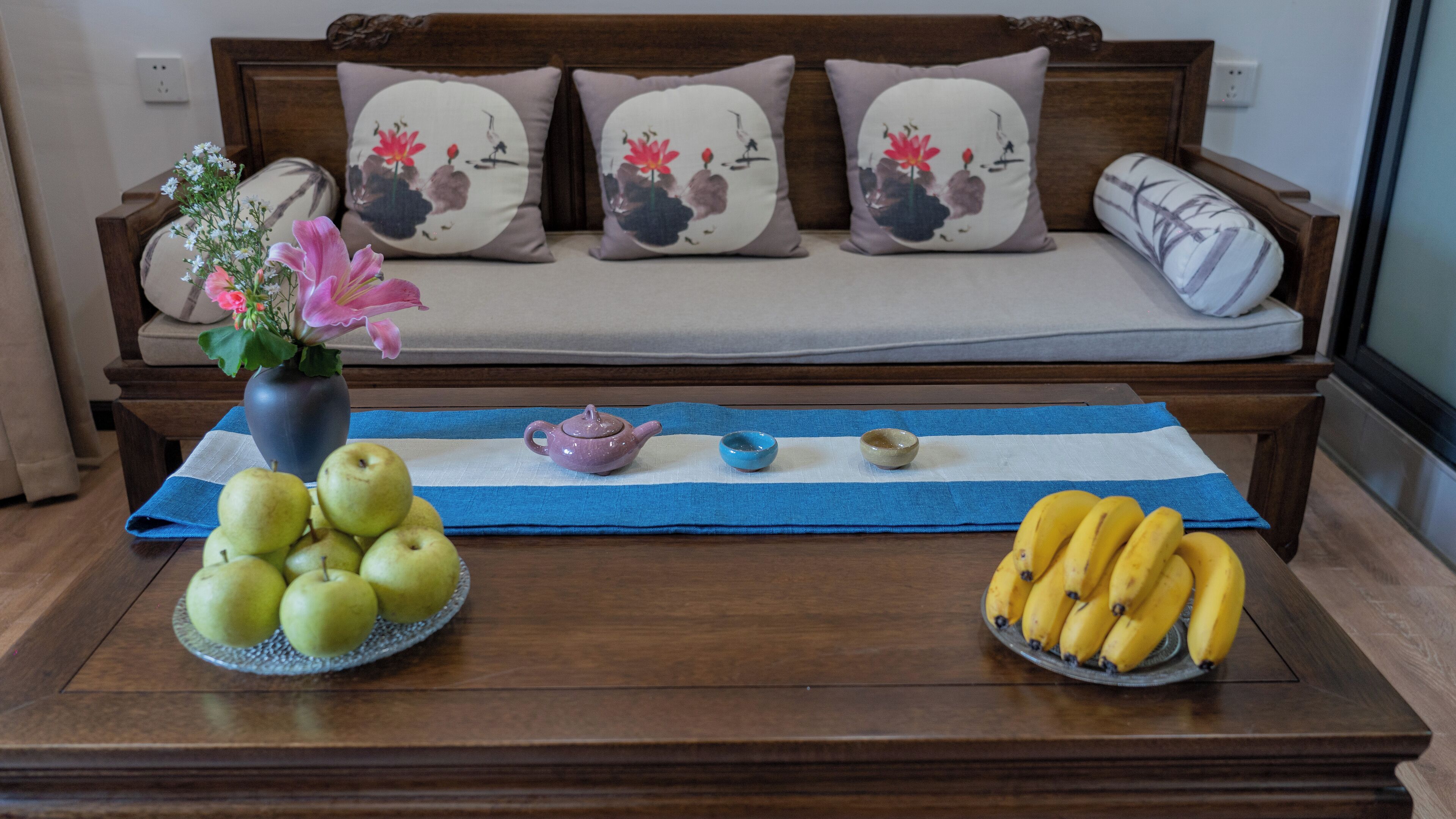 A wooden sofa with floral cushions, a vase with flowers, a teapot, a blue tray with apples and a bowl, and a plate with bananas.