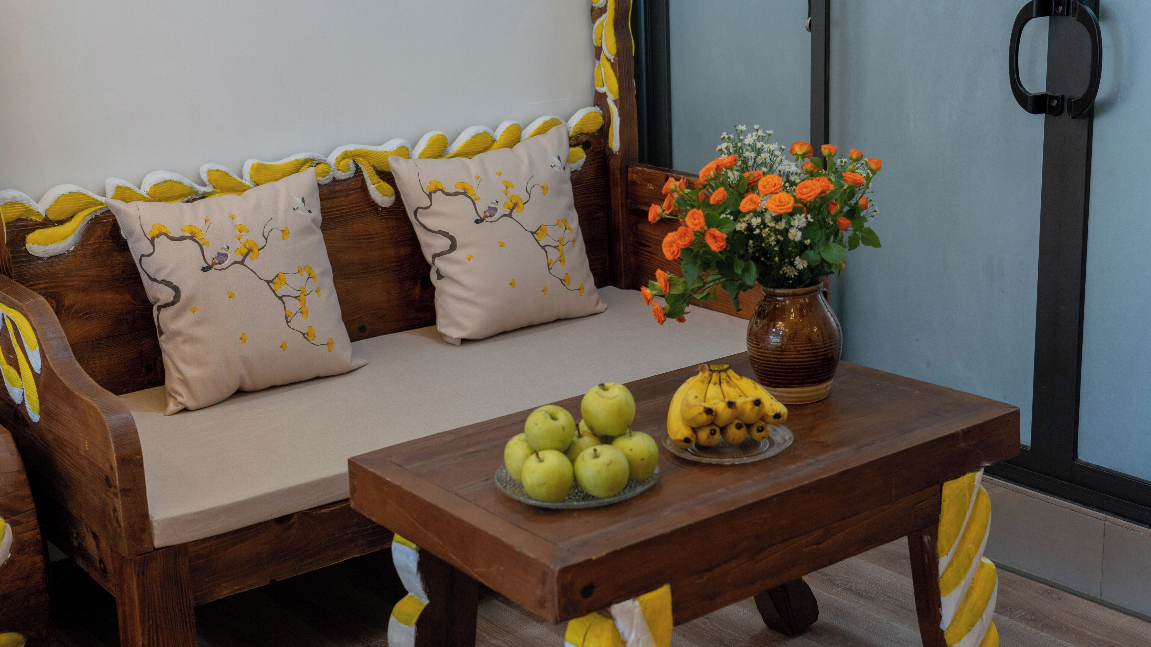 Ein Holzsofa mit gelben und weißen Kissen, ein Couchtisch aus Holz mit Früchten und eine Vase mit Blumen.