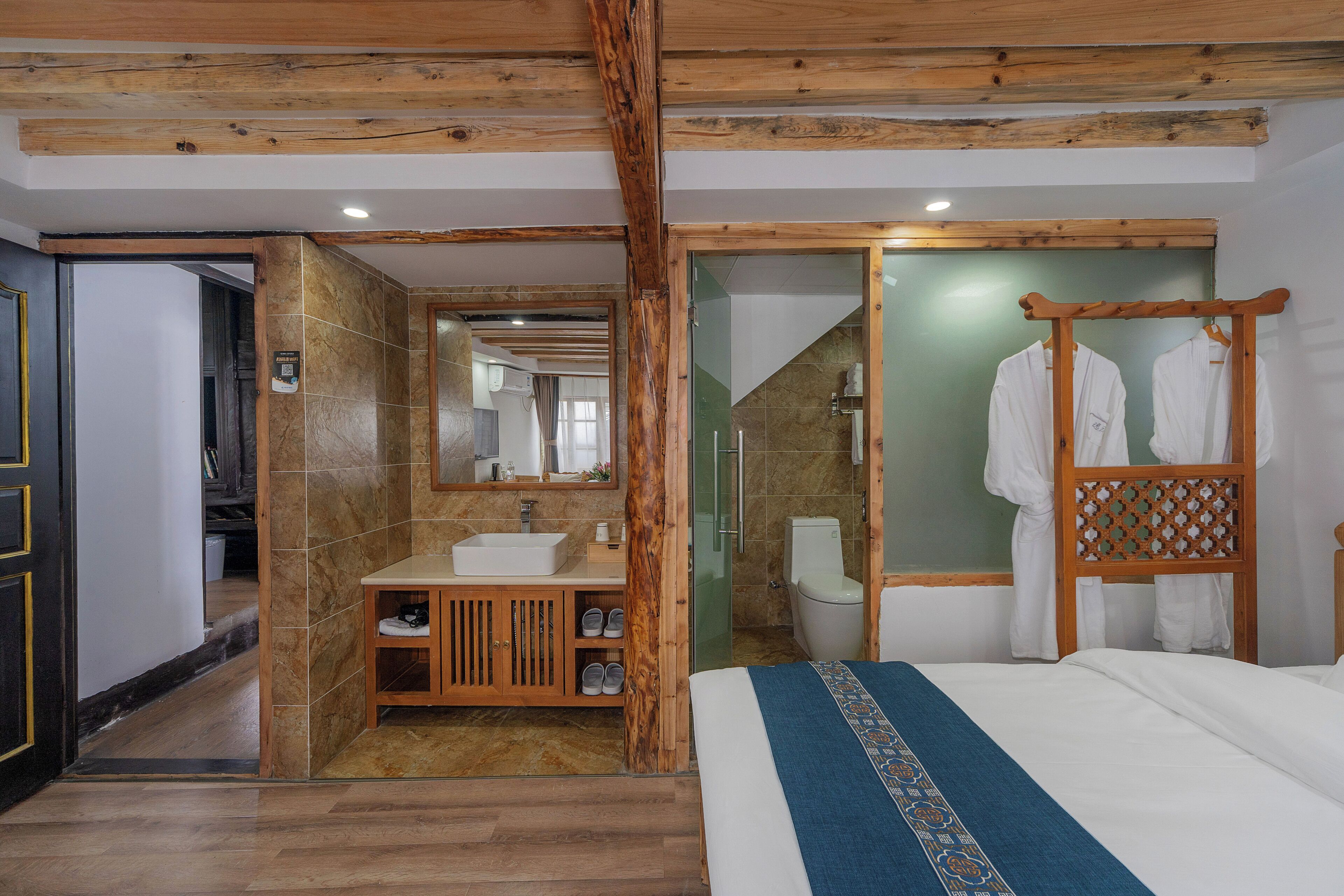 A hotel room with a wooden ceiling, a large bed with a blue bedspread, a wooden vanity with a sink, and a glass shower enclosure.