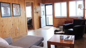 A wooden-paneled room with a sofa, a black leather chair, and a wooden coffee table. There are two framed pictures on the wall and a door leading outside. - Ocean View Cottage, With Access To Tracadie Bay-Minutes From Blooming Point (Mount Stewart)