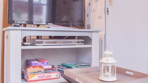 A room with a TV on a white stand, a table with a white lantern, and shelves with books and a Monopoly box. - Gîte De La Voûte in Fécamp with Wi-Fi (Fécamp)