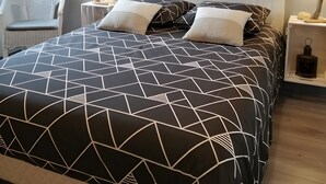 Un dormitorio moderno con una colcha de estampado geométrico, una pintura de paisaje urbano, una silla de mimbre y una mesita de noche con un farol.