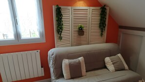 Una habitación con sofá color beige, dos almohadas, un radiador, una ventana y una planta en la pared.