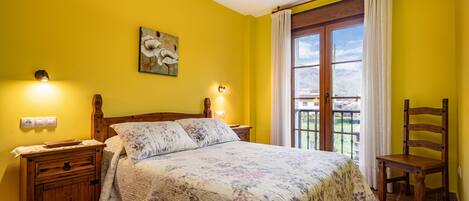 Un dormitorio con una cama, mesitas de noche de madera, una silla, un cuadro y un balcón con cortinas.