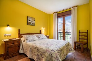 Un dormitorio con una cama, mesitas de noche de madera, una silla, un cuadro y un balcón con cortinas.