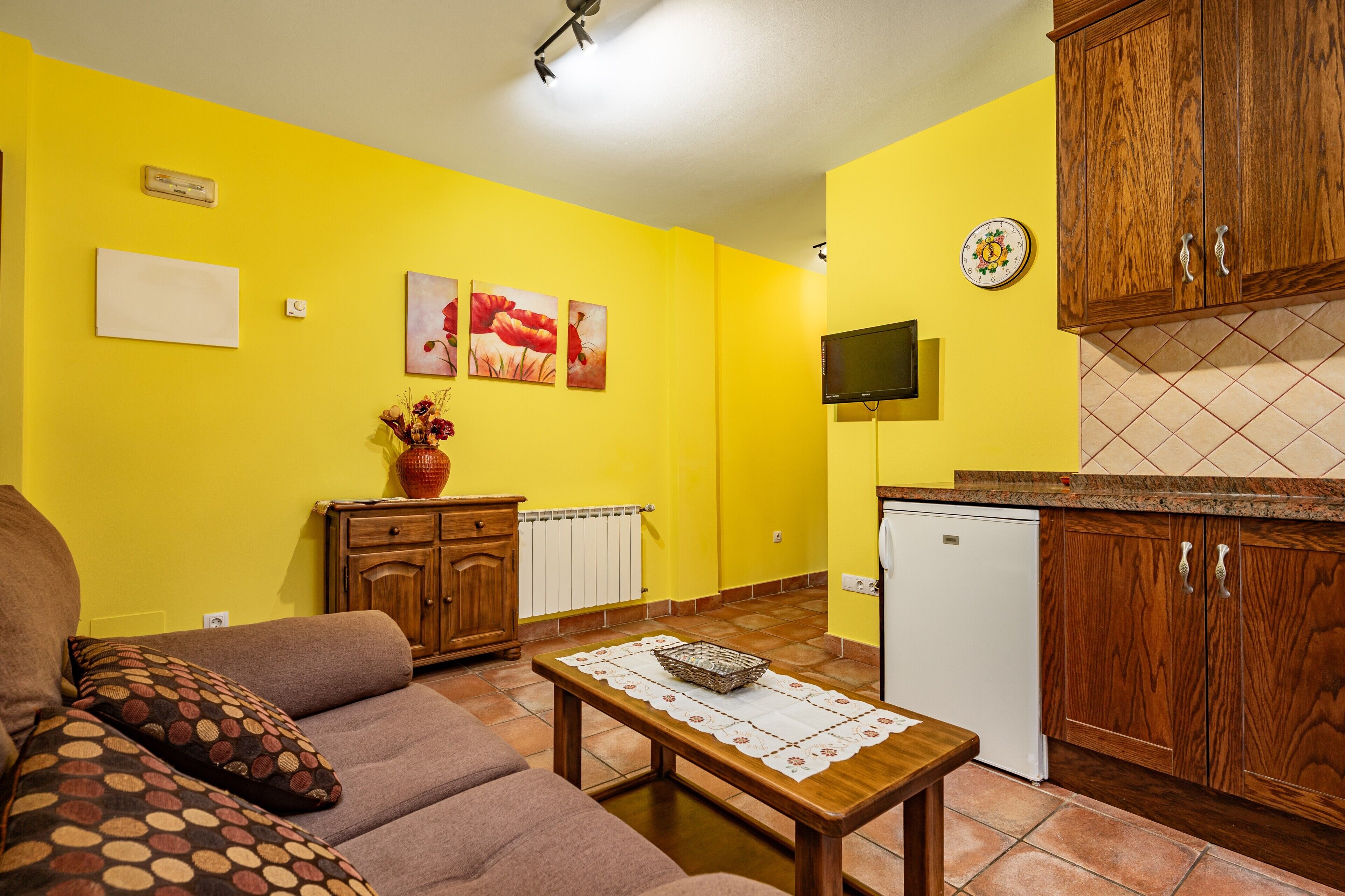 A living room with a sofa, a coffee table, and a kitchen area with wooden cabinets and a tiled backsplash.