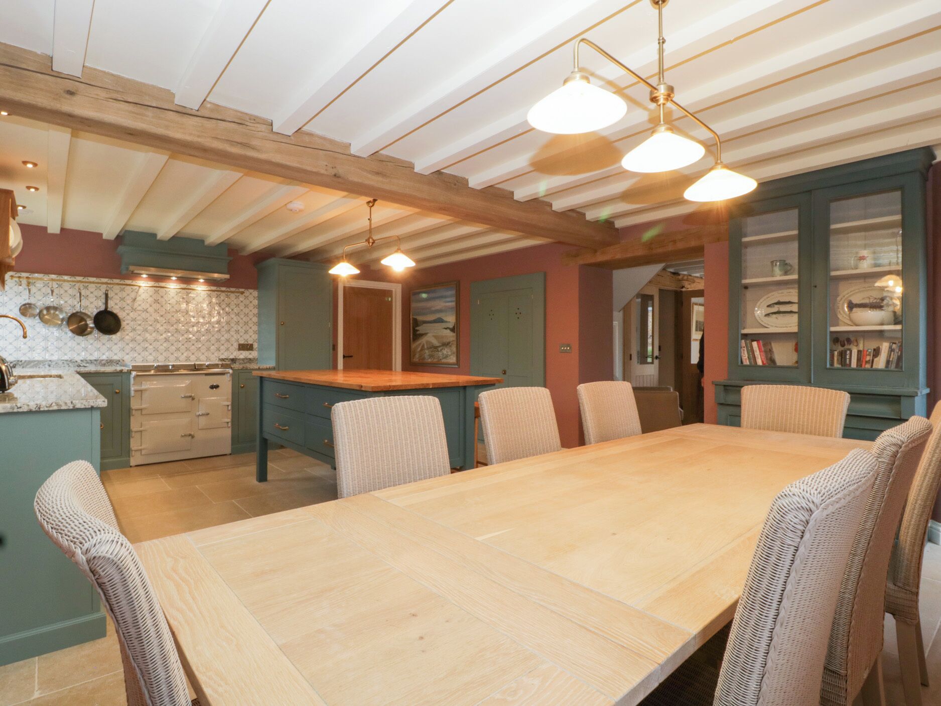 A spacious dining area with a long wooden table and wicker chairs, a kitchen with a stove and cabinets, and a ceiling with exposed beams.