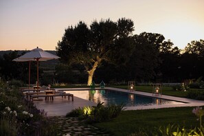 Un área de piscina con sillones de descanso y una mesa, rodeada de follaje y un árbol grande.