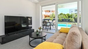Ett modernt vardagsrum med en platt-TV, en soffa med en gul prydnadskudde, ett litet runt bord med en kaktus och utsikt över en swimmingpool och ett utomhusområde med sittplatser.