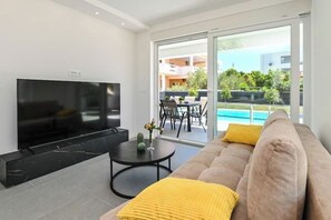 Ein modernes Wohnzimmer mit einem Flachbildfernseher, einem Sofa mit gelber Zierkissen, einem kleinen runden Tisch mit einem Kaktus und Blick auf ein Schwimmbecken sowie eine Außenlounge.