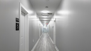 Un pasillo largo y estrecho con paredes blancas y suelo de madera gris. Hay puertas a ambos lados, y un cartel que indica una habitación accesible.