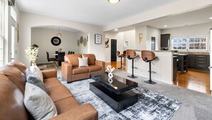 A modern living room with a sofa, coffee table, and bar stools. There is a kitchen area with dark cabinets and a window. - Luxury 4 Bedroom Home Elgin (Elgin)