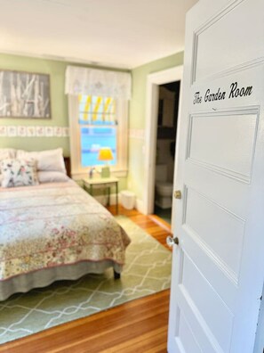 A bedroom with a bed, a nightstand, a lamp, and a window with curtains. The room has a door labeled "The Garden Room." - The Garden Room at the Lafayette Inn (Bar Harbor)