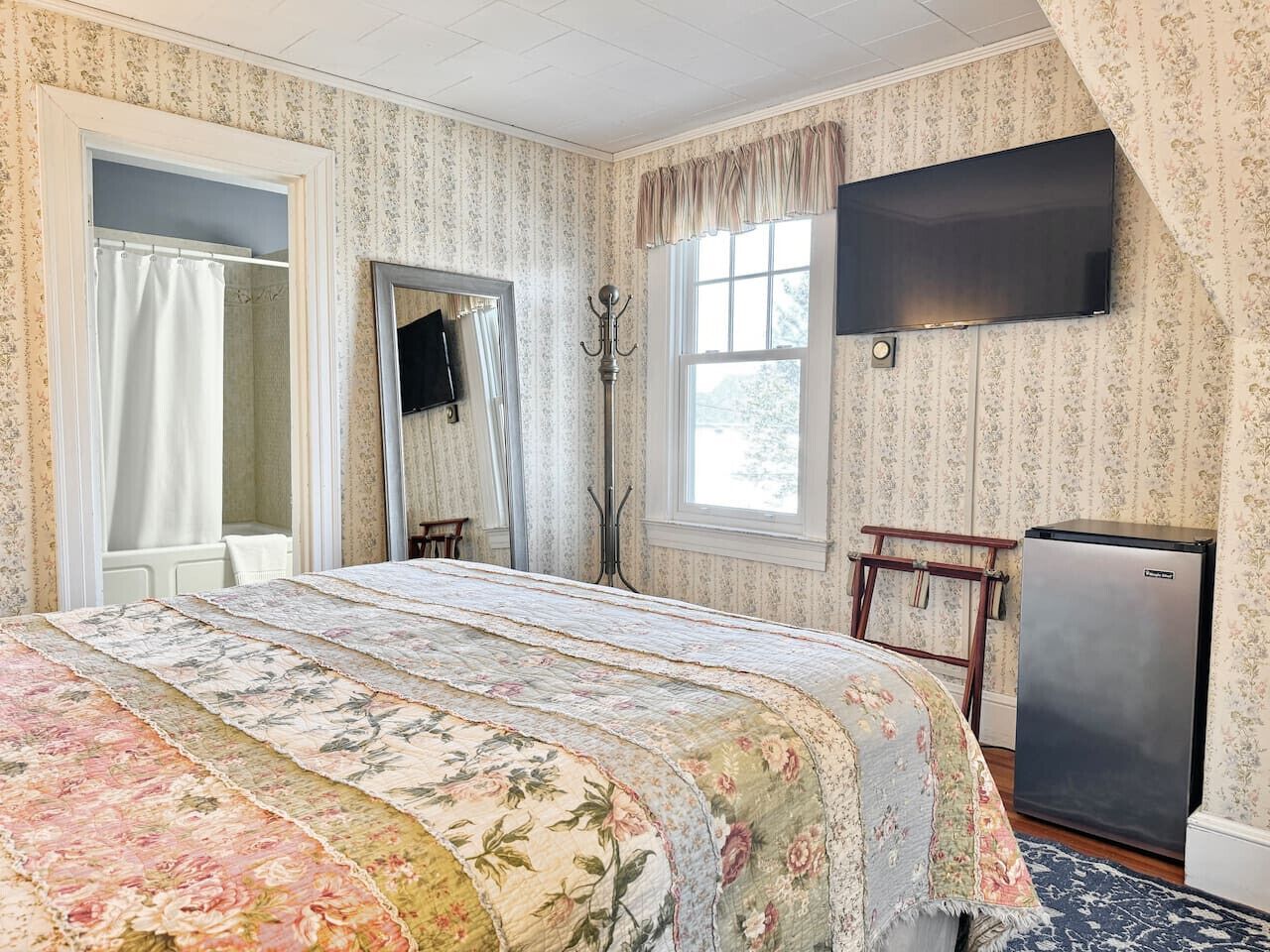 Ein Schlafzimmer mit Bett, Fernseher, Kühlschrank und einem Fenster mit Vorhängen.