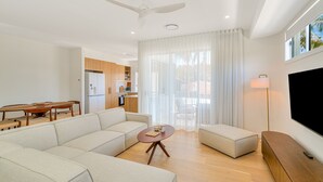 Una sala de estar moderna con sofá, mesa de centro y mueble para televisión. Se observa un área de cocina con alacenas de madera y una mesa de comedor.