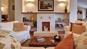 Una sala de estar acogedora con chimenea, dos sofás blancos, una mesa de centro con revistas y estanterías con objetos decorativos.