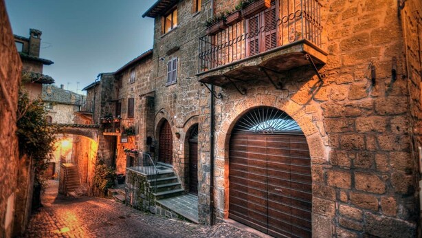 Una estrecha calle de piedra con un edificio que presenta una escalera de madera, arcos en las entradas y un balcón con plantas.