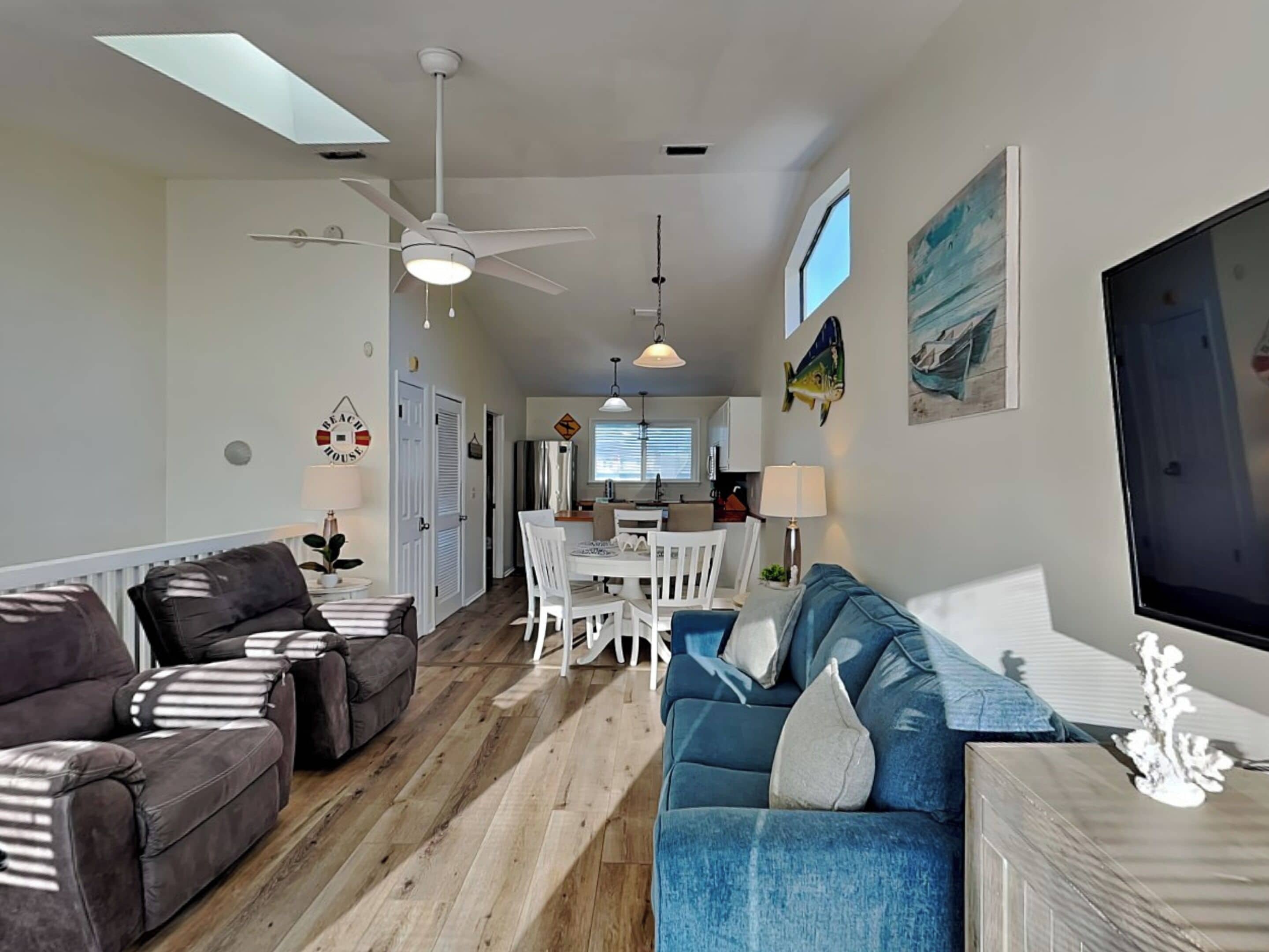 A spacious living area with a wooden floor, a blue sofa, a dark brown sofa, and a dining table with chairs. There is a ceiling fan, a kitchen in the background, and a large window.