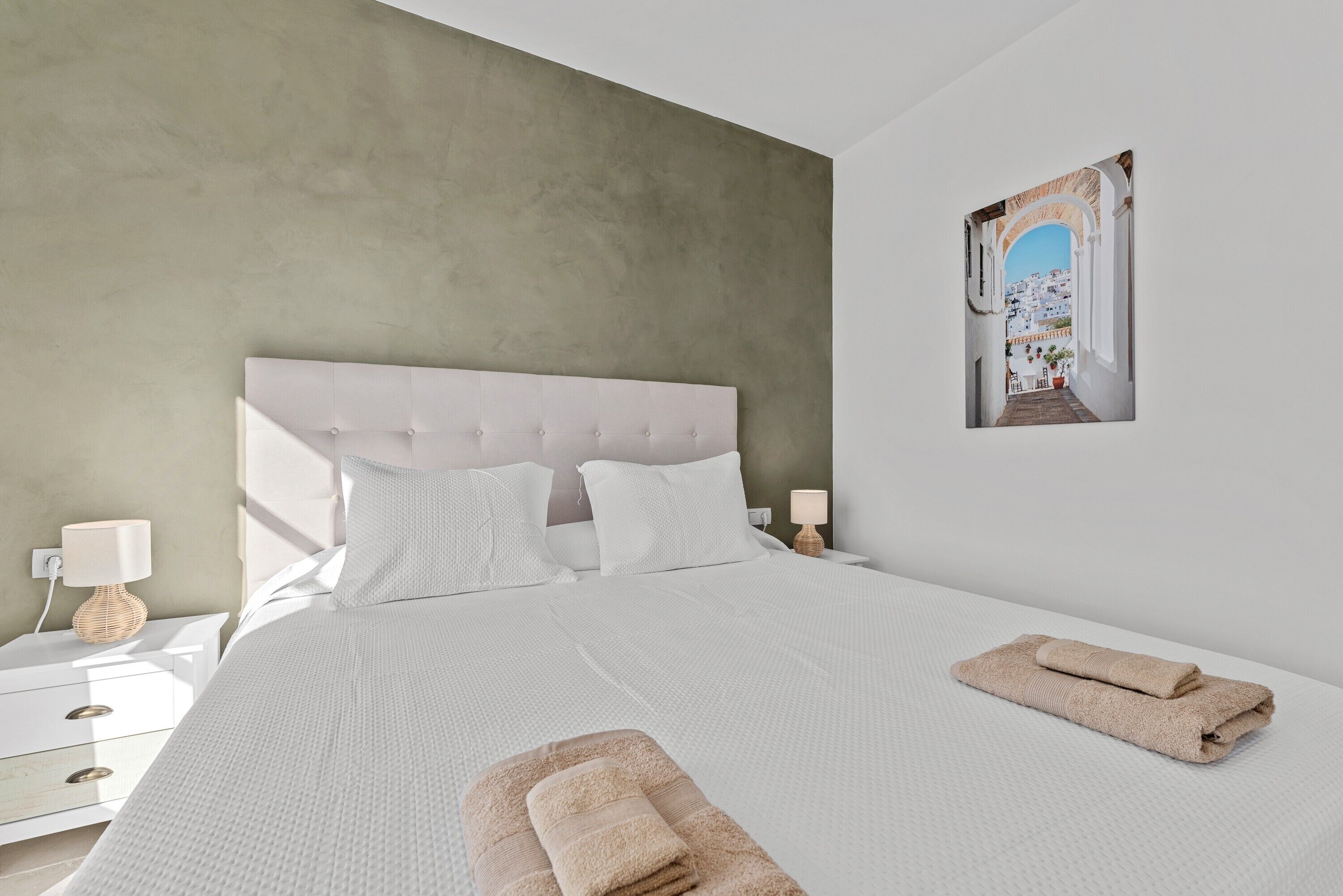 A bedroom with a large bed, white bedding, a headboard, a nightstand with a lamp, and a framed picture on the wall.