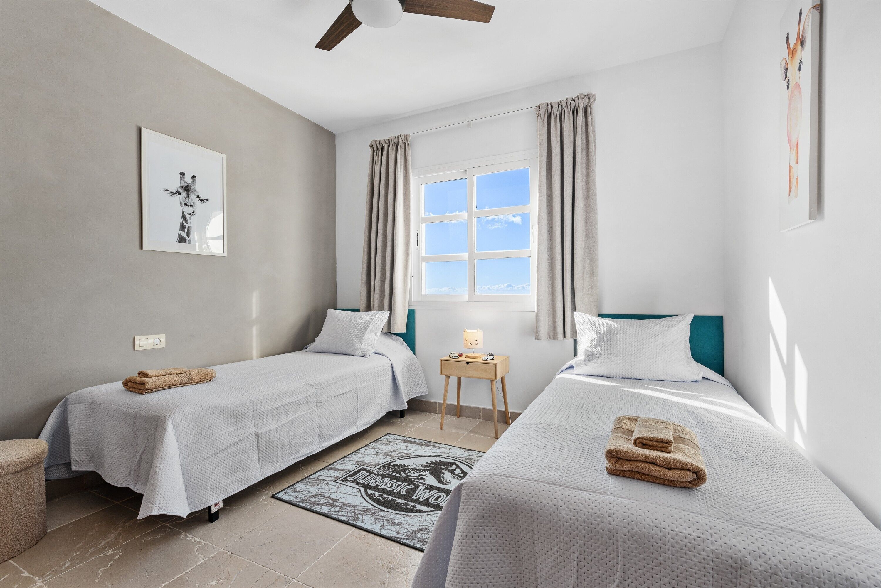 A bedroom with two beds, a window with curtains, a ceiling fan, and a framed picture on the wall.