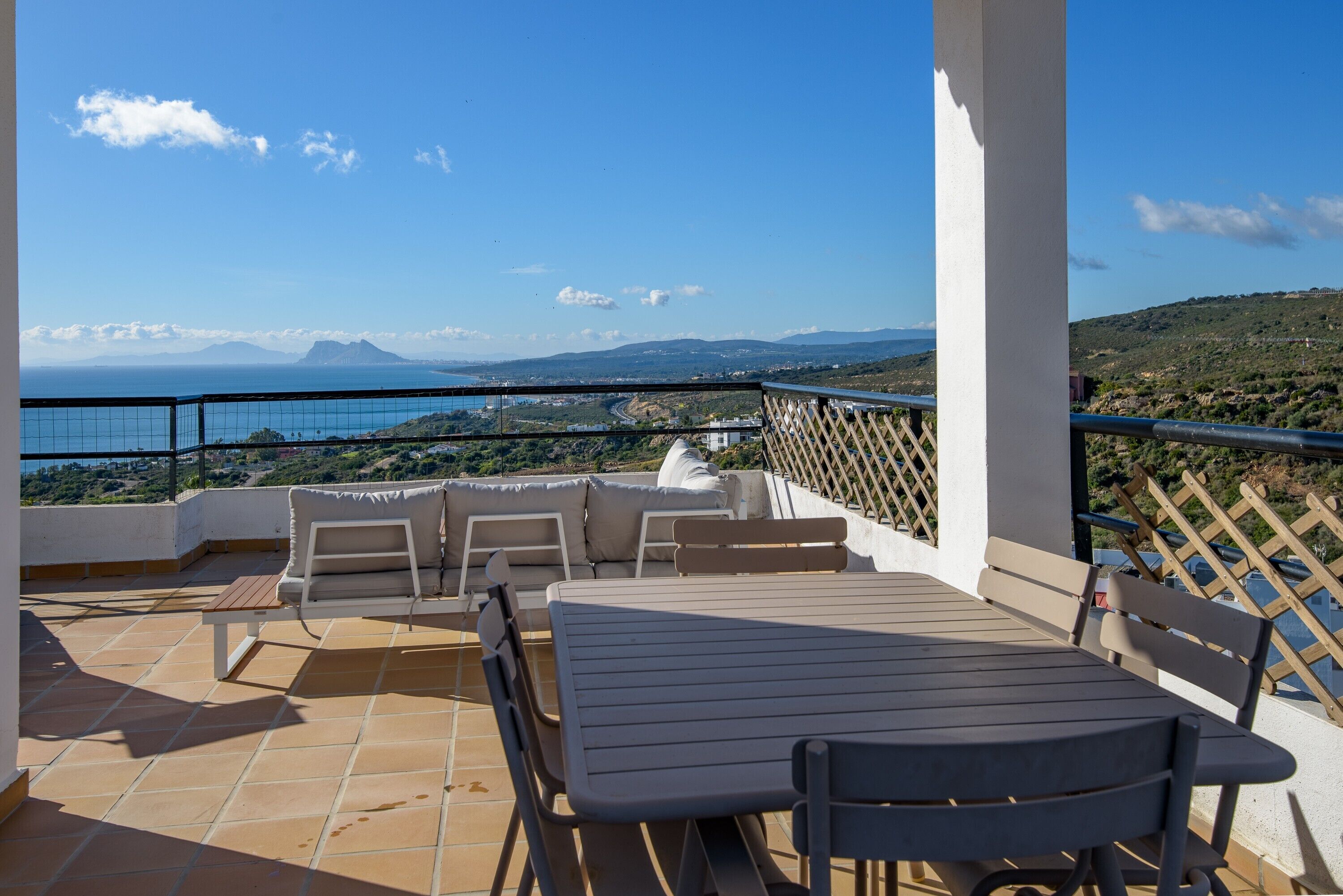 A terrace with a table and chairs, overlooking a coastal landscape with mountains in the distance.