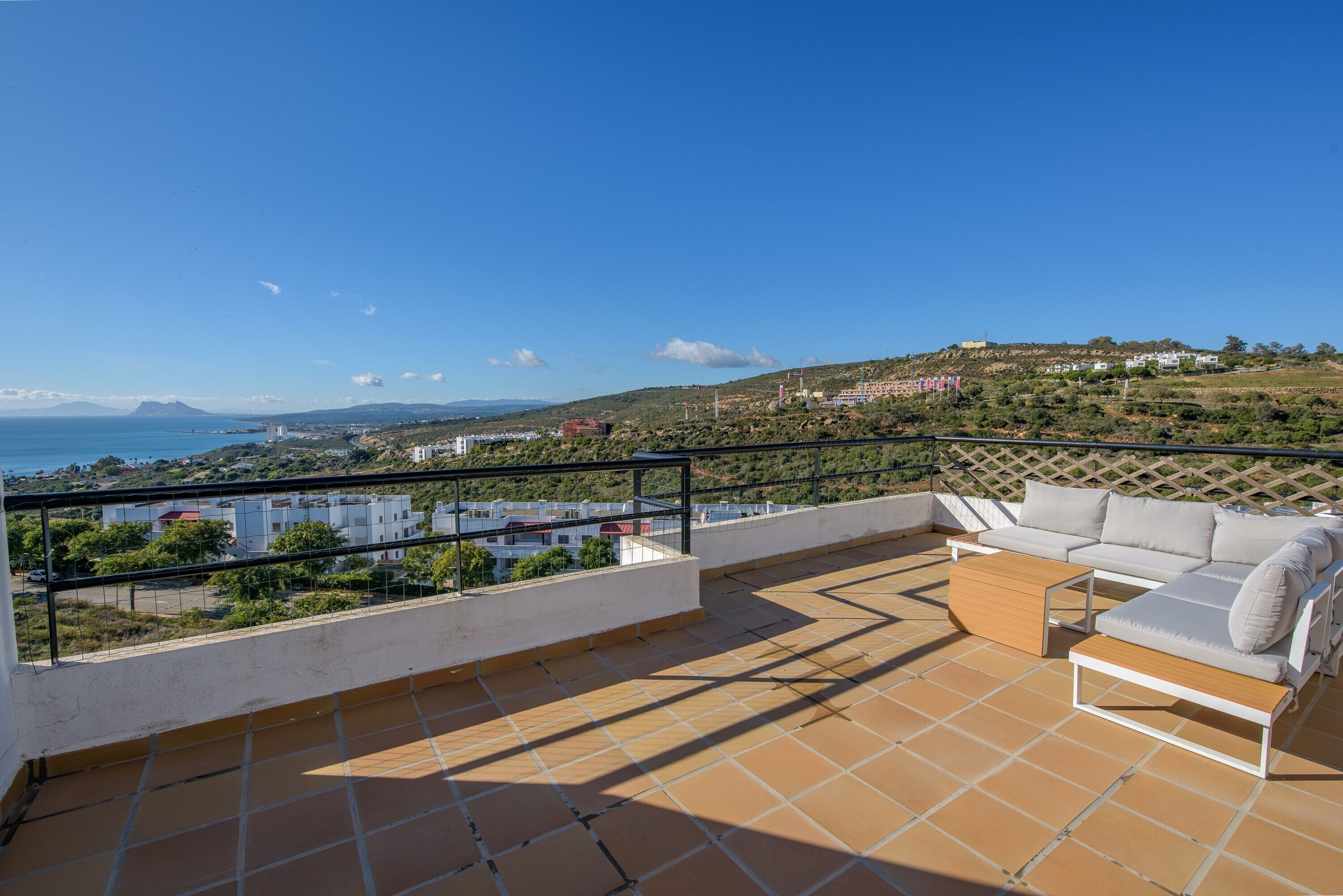 A terrace with a sofa and table, overlooking a coastal landscape with buildings and hills.