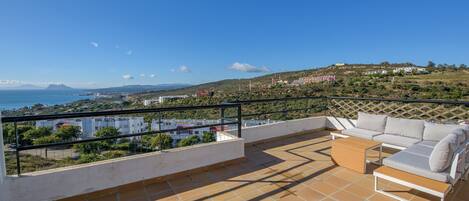 A terrace with a sofa and table, overlooking a coastal landscape with buildings and hills.