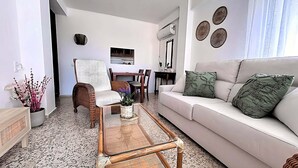 Una sala de estar con un sofá blanco, una silla de mimbre, una mesa de centro de vidrio y arte decorativo en la pared.