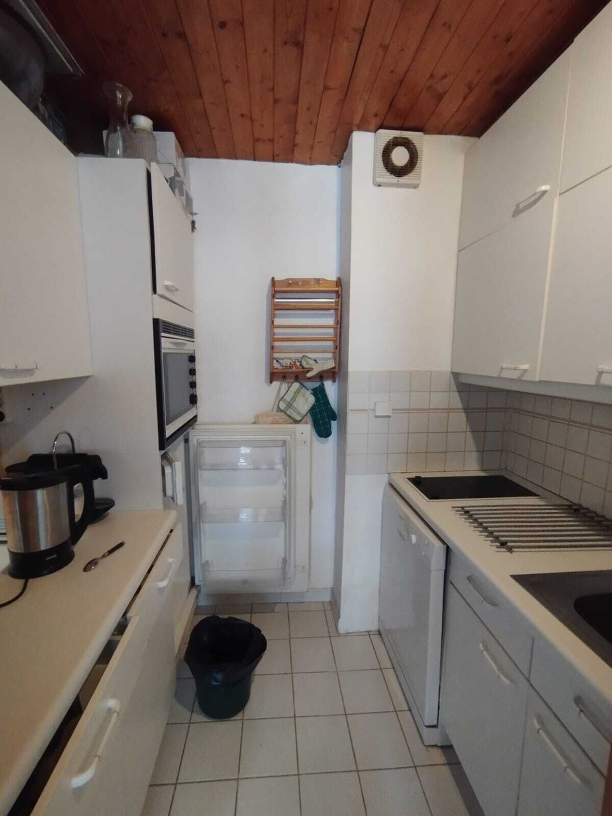 Une cuisine compacte avec des meubles blancs, un four encastré, un four à micro-ondes, une cuisinière et un petit évier. Le plafond est en bois et un porte-serviettes est fixé au mur.