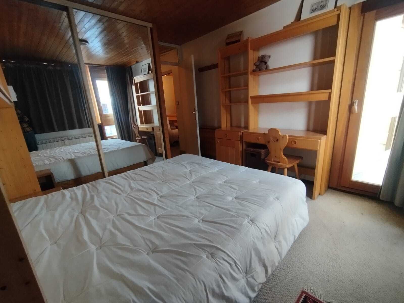 Ein Schlafzimmer mit einem Bett, einem Schreibtisch und einem Stuhl. An der Wand hängt ein Spiegel und es gibt ein Fenster mit Vorhängen.
