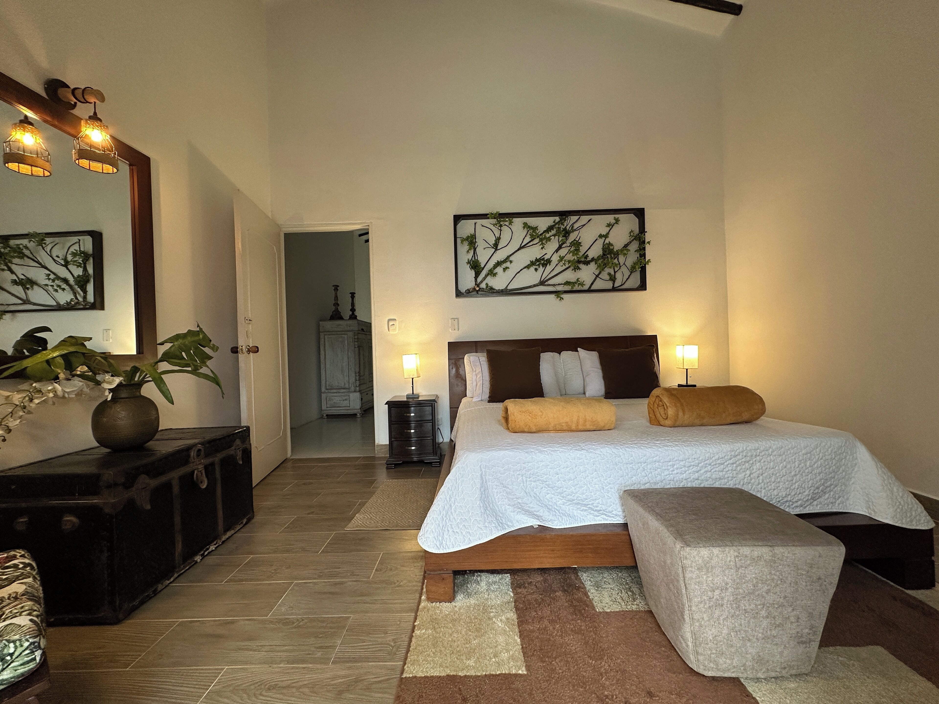 A bedroom with a large bed, bedside lamps, a nightstand, a mirror, a vase with plants, and a painting on the wall.