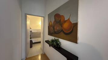 A hallway with a bunk bed in the background, a painting of pears on the wall, and a small plant on a shelf.