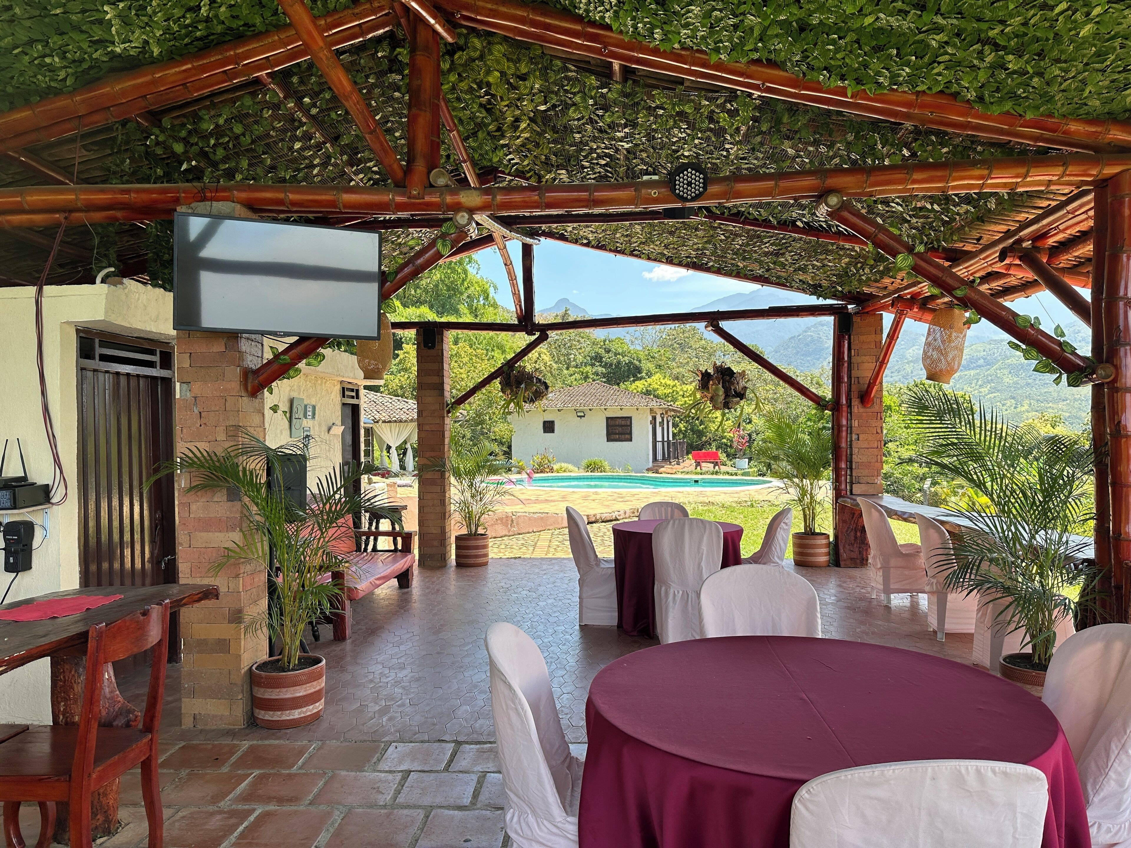 An outdoor covered patio with tables and chairs, a pool, and a view of a house in the background.