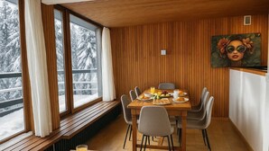 Un comedor con una mesa de madera preparada para el desayuno, ventanas amplias con vistas a árboles cubiertos de nieve y un cuadro en la pared.