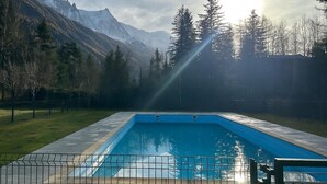 Una piscina rectangular con borde de azulejos, rodeada por una valla metálica, con montañas nevadas y árboles de hoja perenne de fondo, bajo un cielo parcialmente nublado.