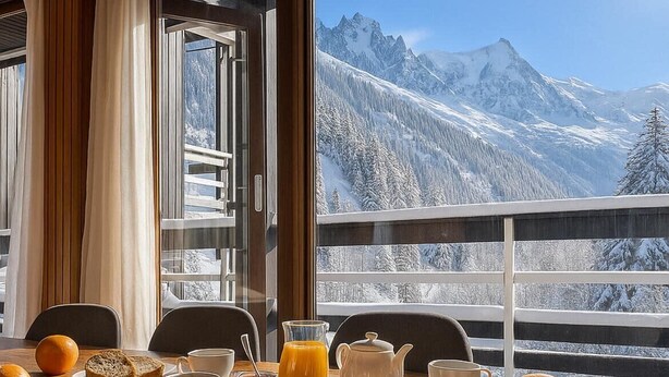 Mesa de desayuno con croissants, pan, zumo de naranja y café, con vistas a un paisaje montañoso nevado.