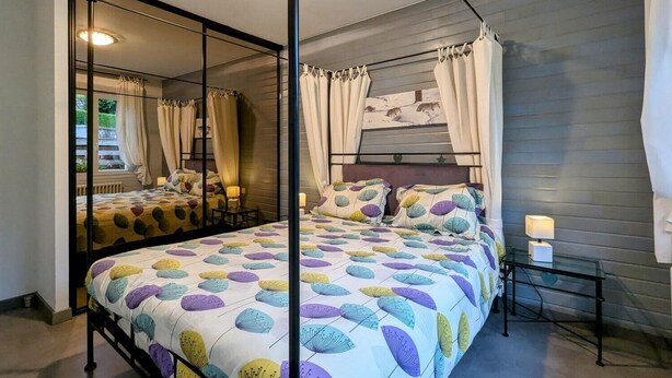 Ein Bett mit Baldachin, eine Bettdecke mit Blätermuster, ein Nachttisch mit Lampe und Blick ins Freie durch ein Fenster mit Vorhängen.