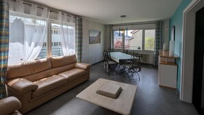 Ein Wohnzimmer mit einer Couch, einem Esstisch mit Stühlen, einem kleinen Teppich und einem Fenster mit Vorhängen.