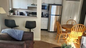 A living room with a sofa, two chairs, a coffee table, and a small table. There is a kitchen in the background with white cabinets, a refrigerator, and a microwave. - Mount Washington’s Mountainside Lodge (Campbell river)