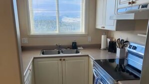 A compact kitchen with white cabinets, a double sink, a stove, and a window with blinds. - Mount Washington’s Mountainside Lodge (Campbell river)