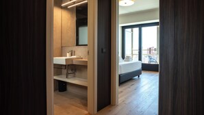 Ein modernes Badezimmer mit einem großen Spiegel, zwei Waschbecken und einer Dusche. Der Raum hat Holzböden und Blick auf einen Balkon mit einem Bett.