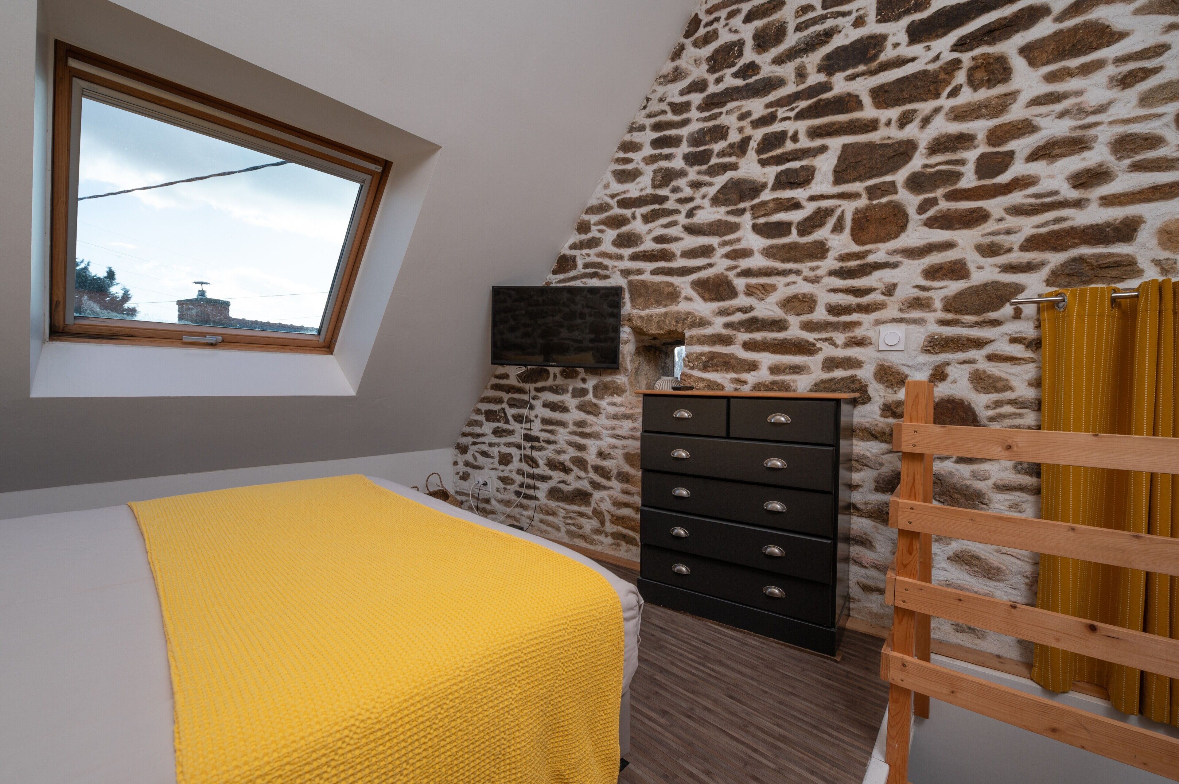 A bedroom with a sloped ceiling, a stone wall, a television, a wooden dresser, and a window with a view.