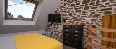 A bedroom with a sloped ceiling, a stone wall, a television, a wooden dresser, and a window with a view.
