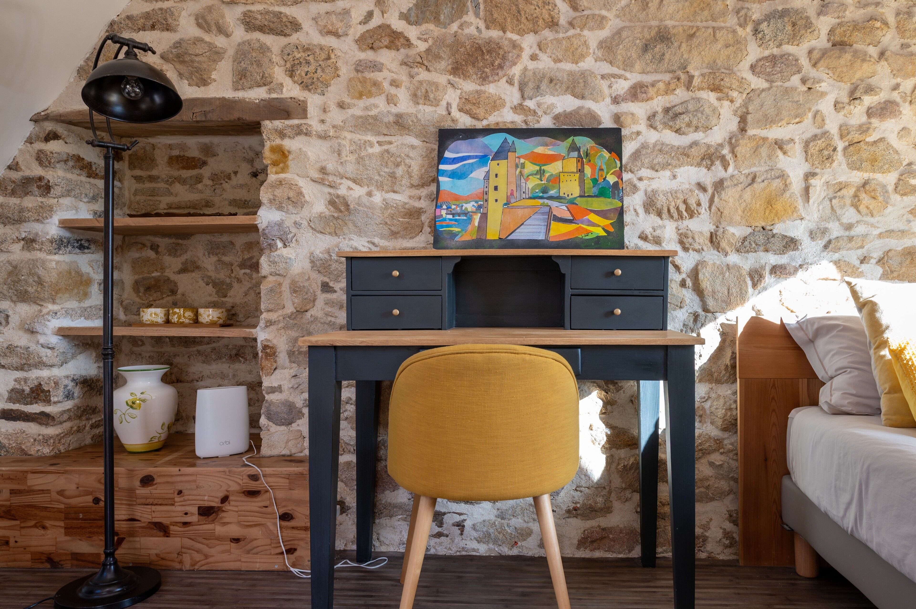 Een kamer met een stenen muur, een bureau met lades, een stoel, een lamp, een bed en een schilderij aan de muur.