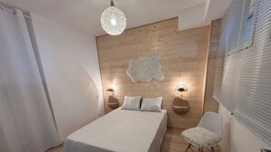 Ein Schlafzimmer mit einem hölzernen Kopfteil, einem Bett mit weißer Bettdecke, zwei Nachttischlampen, einem weißen Sessel und einer Pendelleuchte.