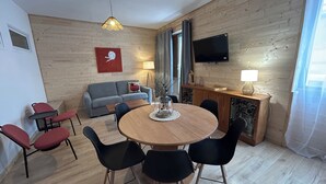 Ein Zimmer mit einem runden Holztisch, umgeben von schwarzen Stühlen, einem grauen Sofa, einem roten Sessel, einem an der Wand montierten Flachbildfernseher und einer Holzkassette mit einer Lampe.