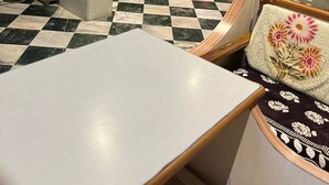 Um chão xadrez, uma mesa branca, uma cadeira de madeira, uma lixeira e um carro estacionado lá fora.