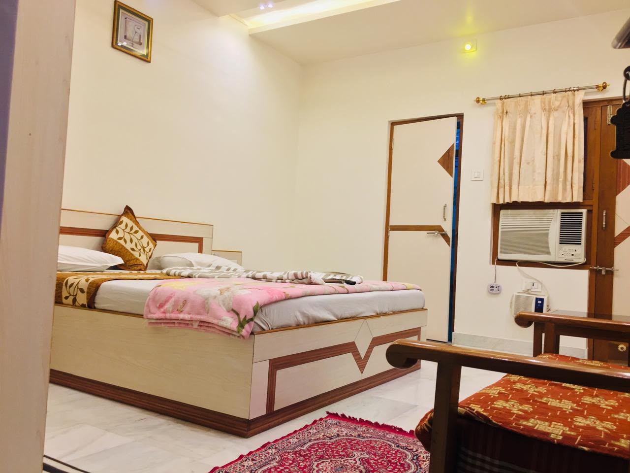 Un dormitorio con cama, mesita de noche, una silla, ventana con aire acondicionado y una puerta.