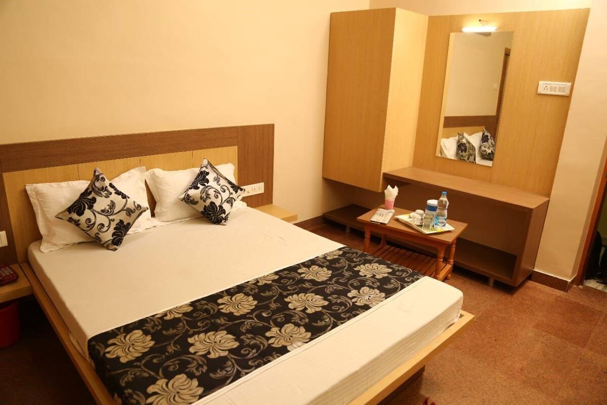 A hotel room with a bed, a bedside table with a bottle and a glass, a mirror, and a small table with a tissue box.