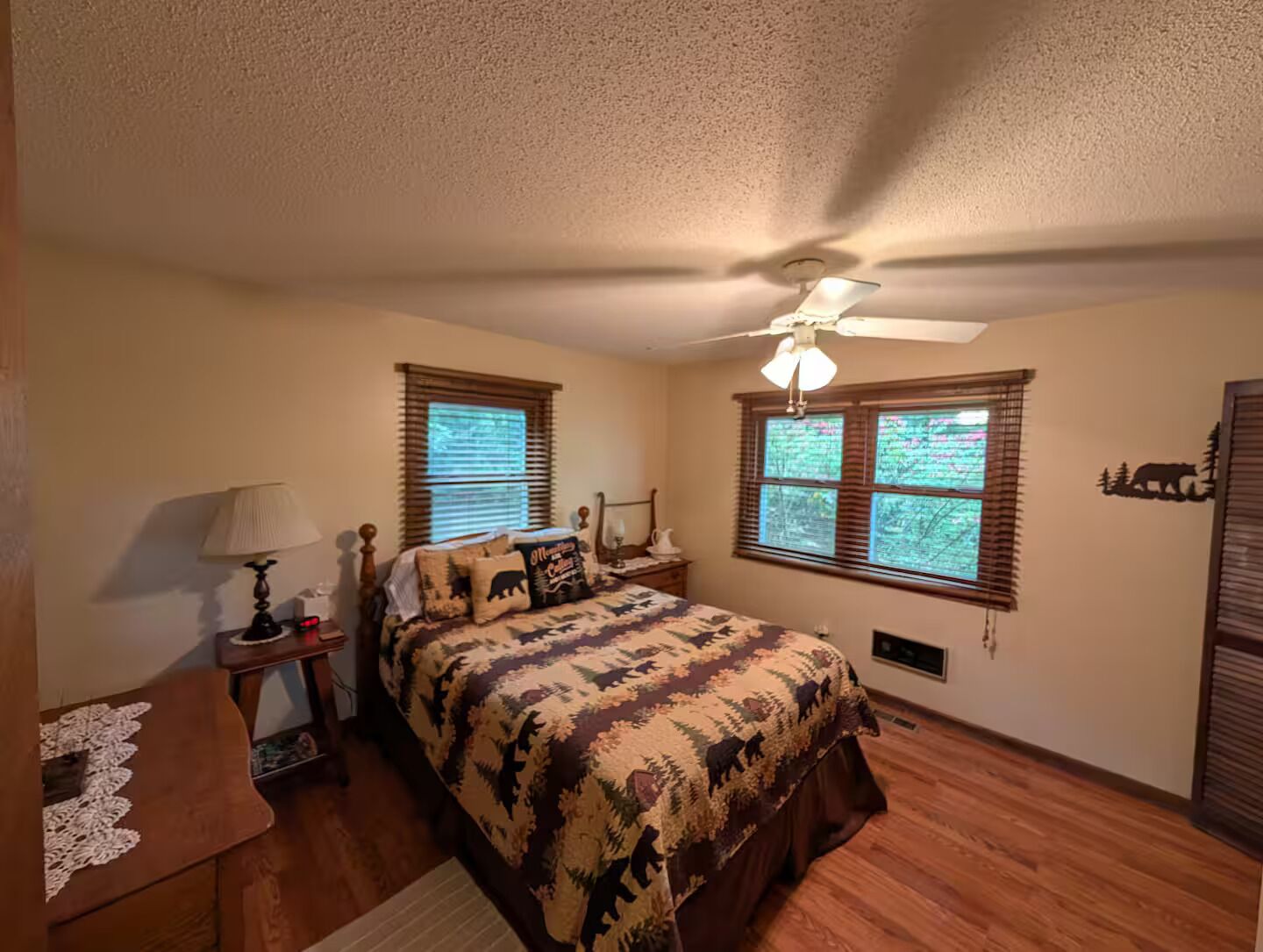 A bedroom with a bed, a nightstand, a lamp, a ceiling fan, and two windows with blinds.